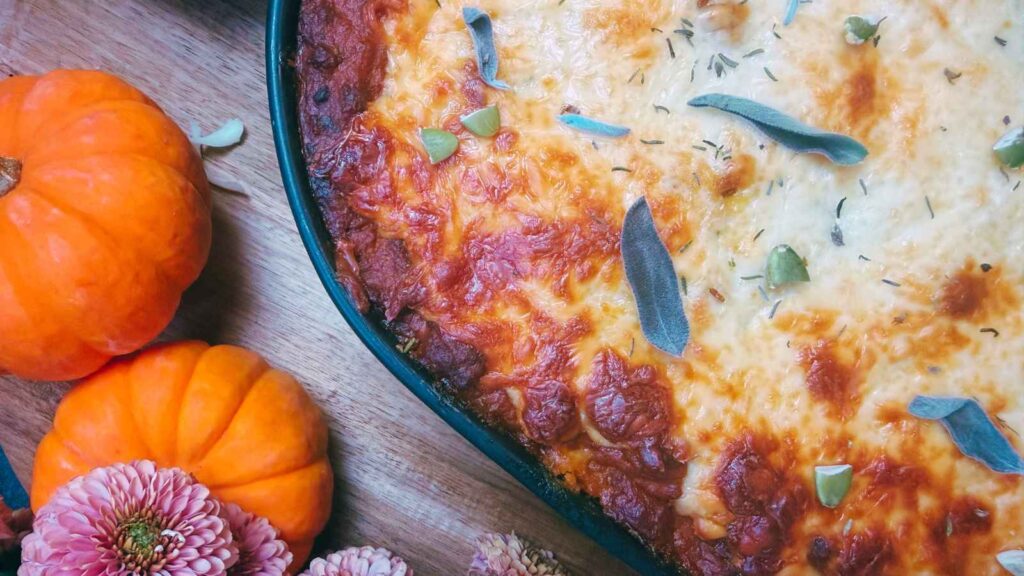 A baked lasagna with browned cheese, sage leaves, and pumpkin seeds on top. There are mini pumpkins and flowers to the side.