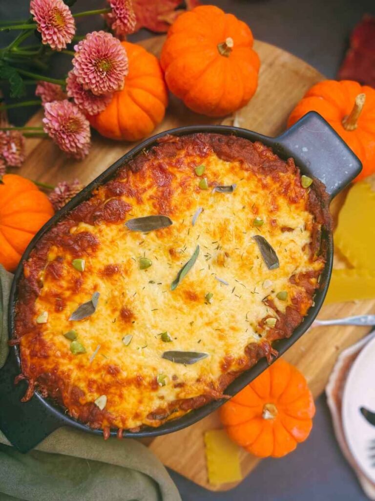 A baked pumpkin lasagna with sage leaves on top. There are mini pumpkins and flowers beside.