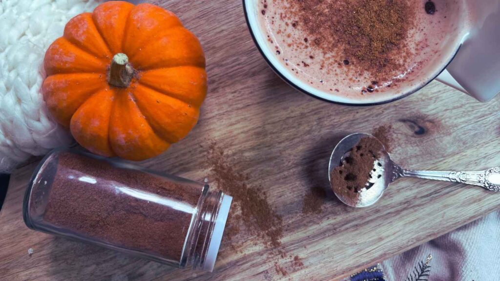 A spice jar tipped on its side. There is a mug of coffee in the background with pumpkin spice on top.