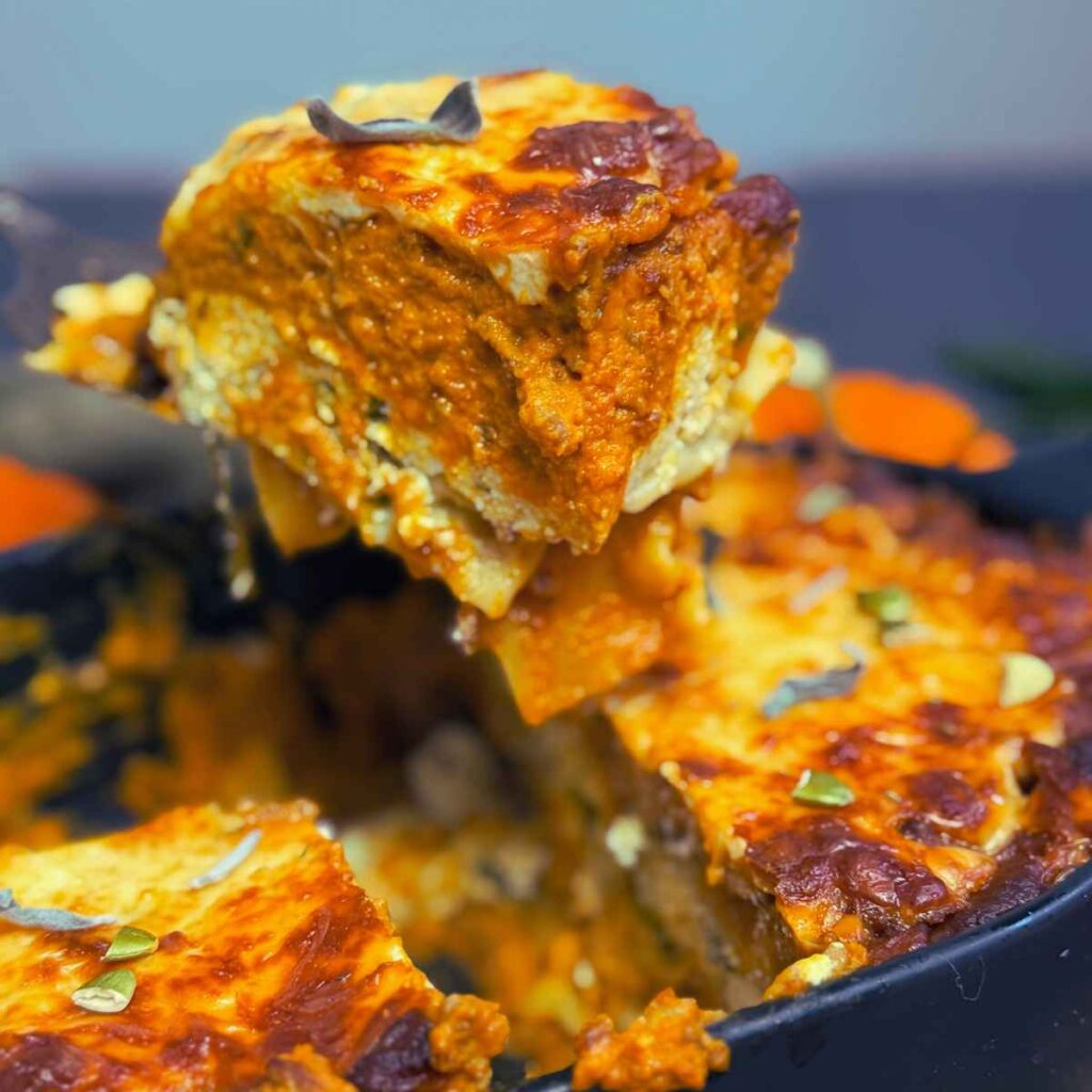 A woman is holding up a portion of pumpkin lasagna. There is more in a black casserole dish below.