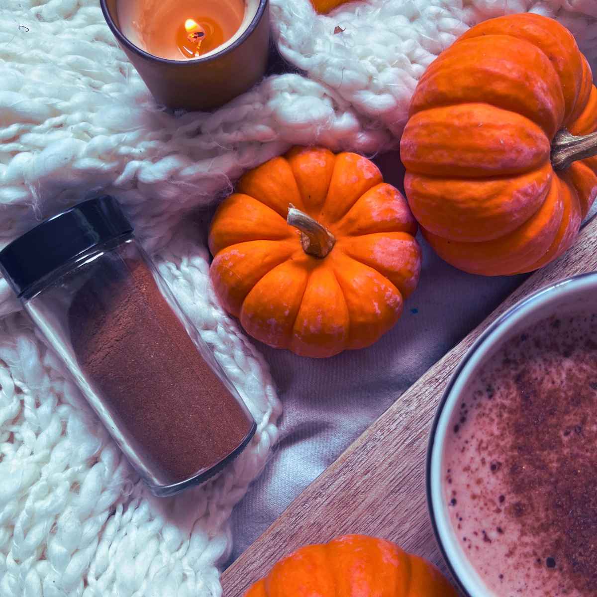 A small jar of pumpkin pie spice on a white knitted scarf with some mini pumpkins, a candle, and a latte with pumpkin pie spice garnish.